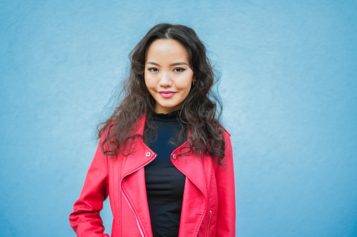 Portrait of happy young asian woman in red leather jacket standing in front of blue background