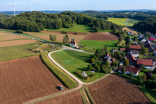 Aerial view of a rural, idyllic landscape with a village among agricultural fields and forests. Wind turbines are visible on the horizon.