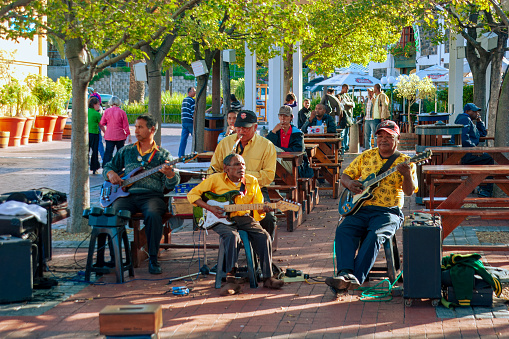 Cape Town, South Africa - A small group of musicians are seen busking by the roadside in the afternoon sunlight. Behind them is some outdoor seating for roadside restaurants.