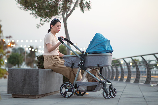 Cute young mother is sitting outside with her newborn baby boy sleeping in a stroller. Woman enjoying in motherhood and spending time with her baby in the fresh, summer air