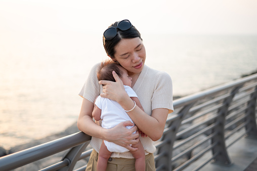 Cute young mother is holding her sleeping newborn baby boy in her arms. A woman enjoying in motherhood and spending time with her baby in the fresh, summer, sea air