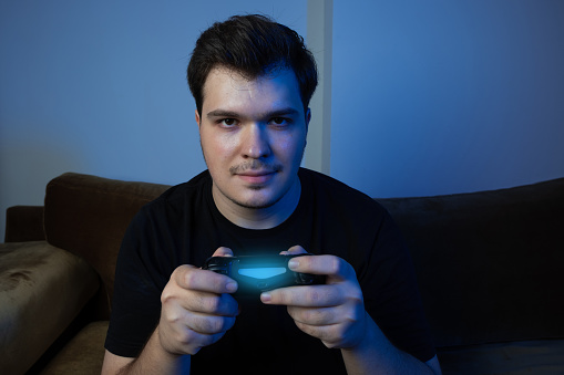 Young man playing console game. Young man smiling and playing video game. He has a joystick in his hand. Blue light dominates the environment. The man is wearing a black t-shirt. He is playing football game. successful gamer