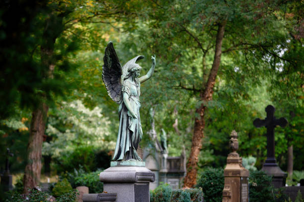 A stone cross in a graveyard, bearing an angel with raised hand. The angel is wearing an ivy head dress. Beckenham Cemetery, Elmers End, London Borough of Bromley.