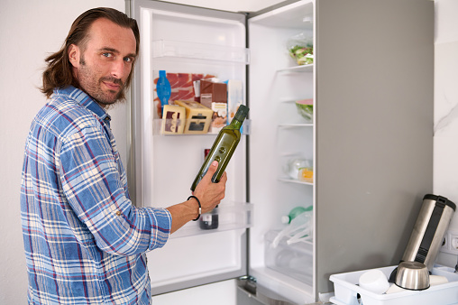 Smiling young adult Caucasian bearded handsome man looking at camera, holding a dark glass bottle with organic virgin olive oil in his hands, standing against an open refrigerator background