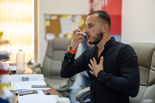 During a busy workday, a young businessman pauses in his office to use his asthma inhaler for necessary relief