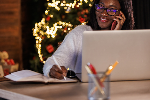 Portrait of joyful young Black entrepreneur sitting at her desk at the office, talking on the phone and writing notes in a note pad while working during Christmas holidays.
