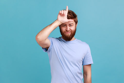 Portrait of handsome you g adult bearded man showing looser gesture holding fingers near forehead, sad because of silly mistake. Indoor studio shot isolated on blue background.