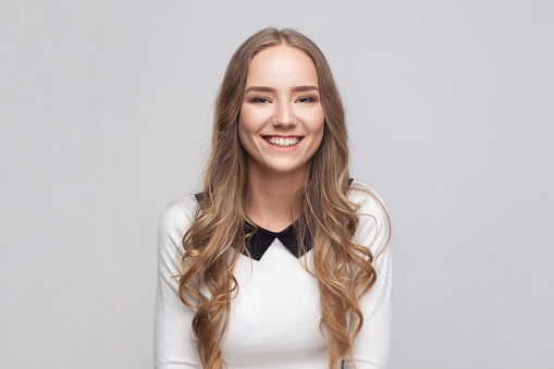 Portrait of charming cute positive attractive woman with wavy blond hair looking at camera with smile, expressing optimism. Indoor studio shot isolated on gray background.