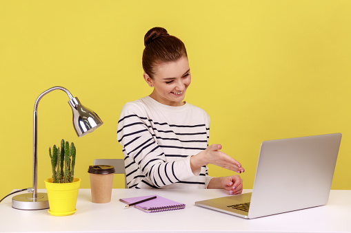 Satisfied woman office manager welcoming client with handshake looking at laptop screen, talking on video call, online conference. Indoor studio studio shot isolated on yellow background.