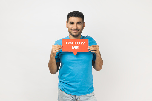 Portrait of joyful satisfied handsome unshaven man wearing blue T- shirt standing holding card with follow me inscription, looking at camera. Indoor studio shot isolated on gray background.