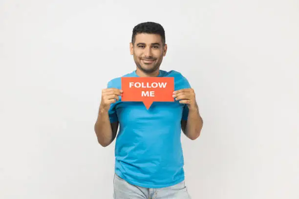 Photo of Joyful satisfied handsome man standing holding card with follow me inscription, looking at camera.