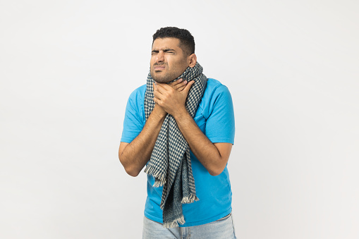Portrait of unhealthy unshaven man wearing blue T- shirt and wrapped in warm scarf standing touching neck, having sore throat. Indoor studio shot isolated on gray background.