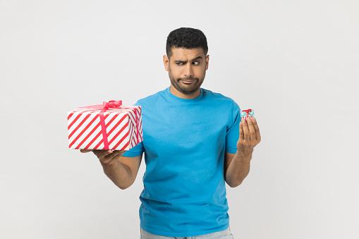 Portrait of pensive confused unshaven man wearing blue T- shirt standing holding two present boxes, little and big, thinking what to choose. Indoor studio shot isolated on gray background.