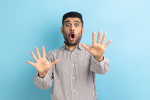 I'm afraid. Portrait of shocked scared bearded businessman screaming in horror and fear, keeping hands raised to defend himself, wearing striped shirt. Indoor studio shot isolated on blue background.