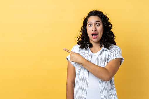 Portrait of amazed shocked beautiful woman with dark wavy hair standing pointing aside with finger, showing copy space for advertisement. Indoor studio shot isolated on yellow background.