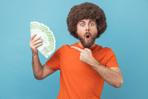 Portrait of surprised amazed man with Afro hairstyle wearing orange T-shirt holding euro banknotes, being shocked to win lottery. Indoor studio shot isolated on blue background.