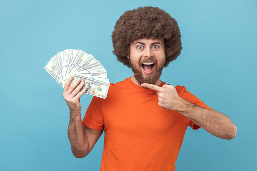 Portrait of excited positive man with Afro hairstyle wearing orange T-shirt holding dollar banknotes, being happy to win lottery. Indoor studio shot isolated on blue background.