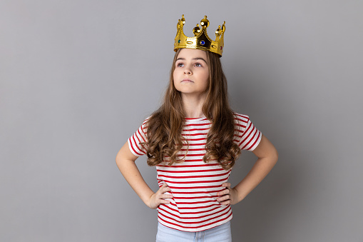 Portrait of confident little girl princess wearing striped T-shirt keeping hands on hips, self-motivation and dreams to be best. Indoor studio shot isolated on gray background.