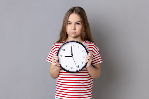 Portrait of serious little girl wearing striped T-shirt holding big wall clock, looking at camera with unpleasant emotions, time to go. Indoor studio shot isolated on gray background.