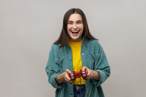 Excited woman holding gamepad, winning video games competition, looking at camera with happy expression, wearing casual style jacket. Indoor studio shot isolated on gray background.
