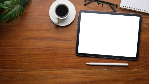 istock Flat lay, top view of digital tablet with white screen, coffee cup, glasses and notepad on wooden working desk. 1739875235