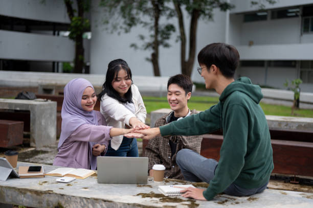 A group of cheerful, diverse Asian university students is putting their hands together at a table in a campus park, encouraging and supporting each other as a team.