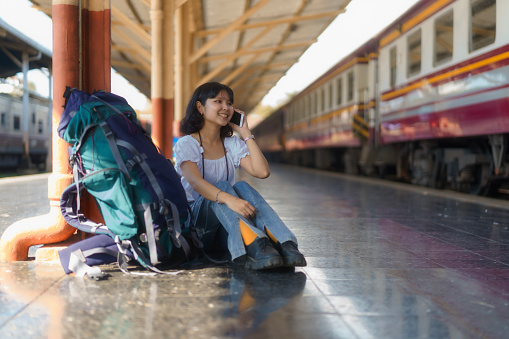 An Asian female tourist is using a mobile phone and a backpack on the platform of a train station.
