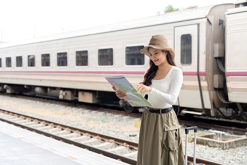 Happy asian woman traveler using map choose where to travel with luggage at train station, summer vacation travel concept.