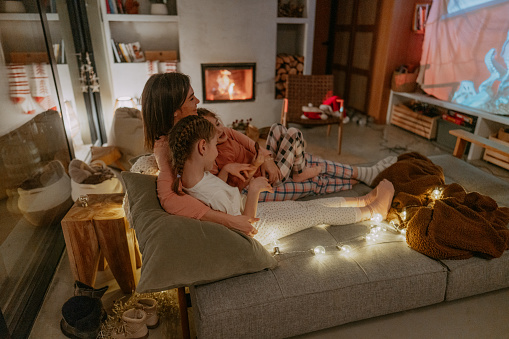 Photo of a two young girls and their mom who enjoy watching movies on Christmas Eve at home. They are watching videos on an improvised projector screen.