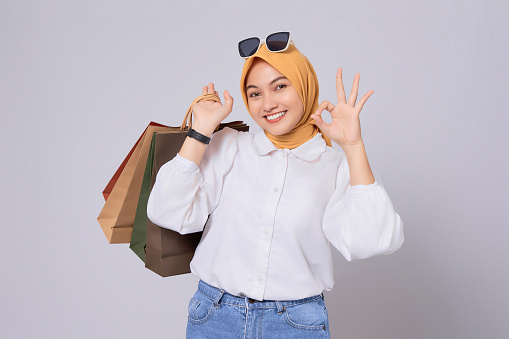 Beautiful smiling young Asian shopaholic Woman carrying shopping bags and showing okay sign isolated on white background. online mobile shopping concept