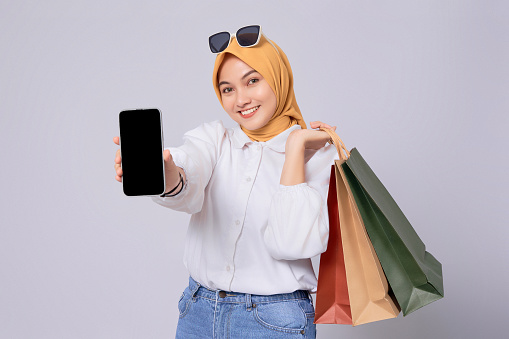 Beautiful smiling young Asian Muslim Woman showing mobile phone with a blank screen and holding shopping bags isolated on white background. online mobile shopping concept