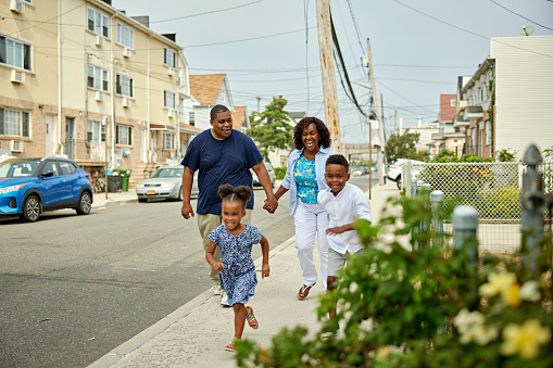 Mature Black couple holding hands and laughing as they follow their speedy 4 and 7 year old children, grinning as they enjoy weekend fun in the neighborhood.