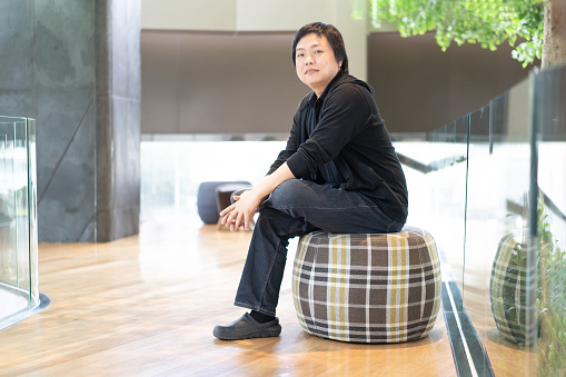 In the contemporary hall of a modern building, an Asian man dressed in black attire finds comfort as he sits on a circular sofa.