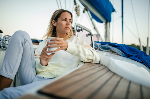 Beautiful young adult woman relaxing on sailboat deck and enjoying coffee or tea. She is wearing warm clothes in calm, autumn morning on sailing off-season vacation, Ionian sea, Kefalonia island, Greece