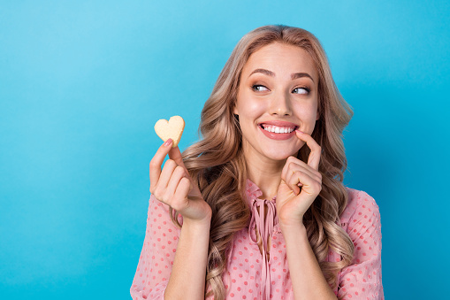 Photo of funny pretty woman with wavy hairdo dressed pink clothes look at heart shape cookie in fingers isolated on blue color background.