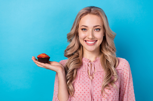 Photo of good mood adorable woman with wavy hairdo dressed pink clothes arm holding sweet muffin isolated on blue color background.