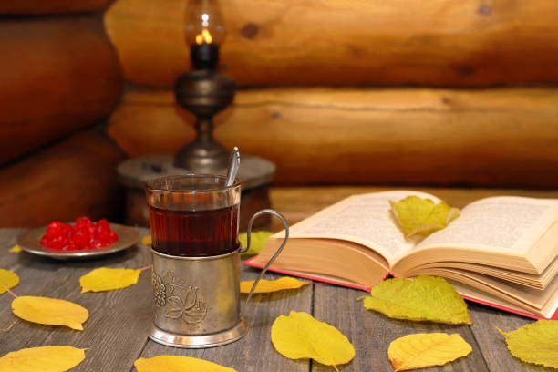 Autumn tea party in retro style. On boards table among autumn foliage is glass of tea in iron holder next to iron plate with dried cherry berries and book on background of  kerosene lantern standing on wooden barrel near log wall. tea party horizontal nobody indoors stock pictures, royalty-free photos & images
