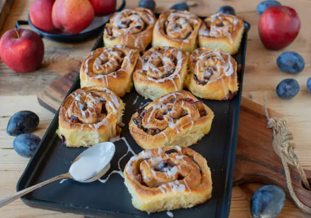 Delicious homemade sticky cinnamon rolls with plum filling. Sweet autumn pastry. Fresh glazed with sugar glaze and ready to eat on a baking sheet.