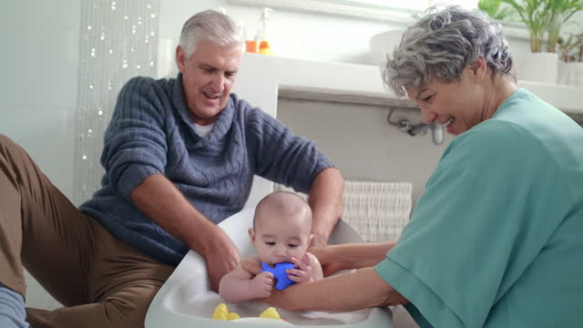 Baby, bath and happy grandparents in home, cleaning and hygiene for health. Elderly man, woman and washing child body with soap, play in water and rubber duck for care, bonding and family in bathroom
