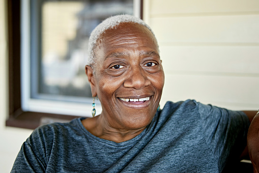 Close view of woman with short gray hair wearing summertime clothing and smiling at camera on front porch.