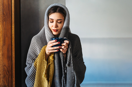 Freezing woman covered with a cozy blanket. Female wrapped in warm blanket, drinking hot drink. Cold flu concept.