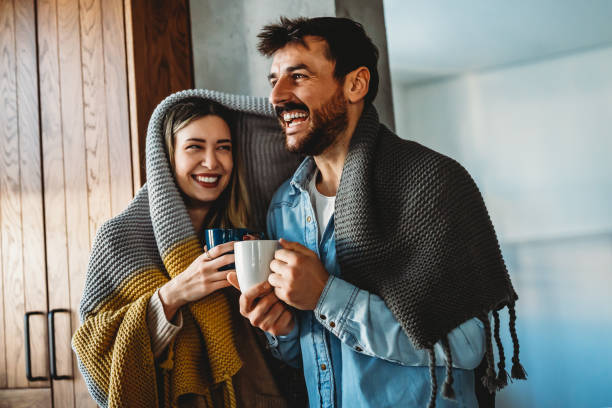 istock Happy young couple with hot drinks cuddling under warm blanket at home 1740368778