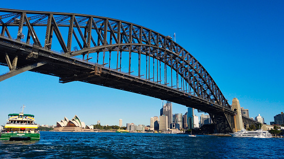 Milsons Point, Sydney, Australia - June 09 2019 : The huge span of the bridge with the Bungaree Ferry underneath it apart to depart from the jetty on a warm afternoon.