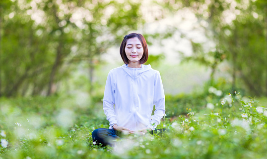 Asian woman is relaxingly practicing meditation yoga in the forest full of daisy flower in summer to attain happiness from inner peace wisdom with morning light for healthy mind and soul