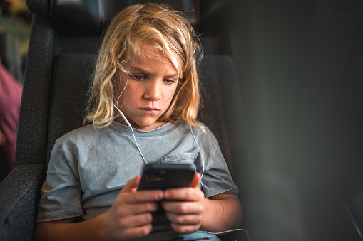 Caucasian boy, with blond hair, keeps entertained during his train journey with a smartphone and earphones.