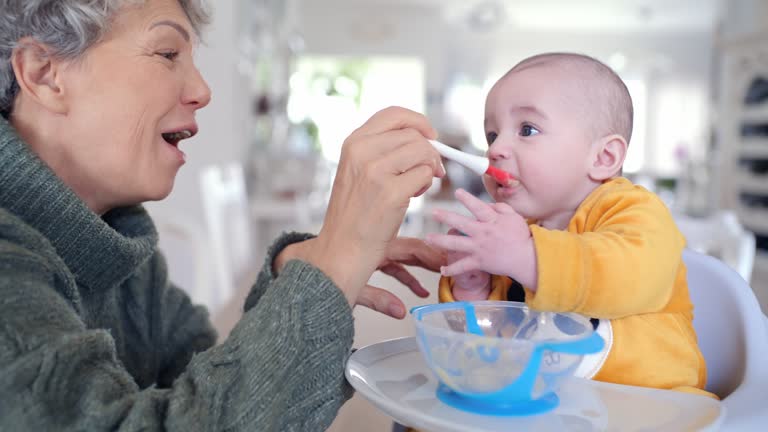 Happy grandmother feeding baby in home, love and family bonding in kitchen for morning breakfast. Smile, food and old woman with hungry child eating lunch in apartment, babysitting and fun at table.