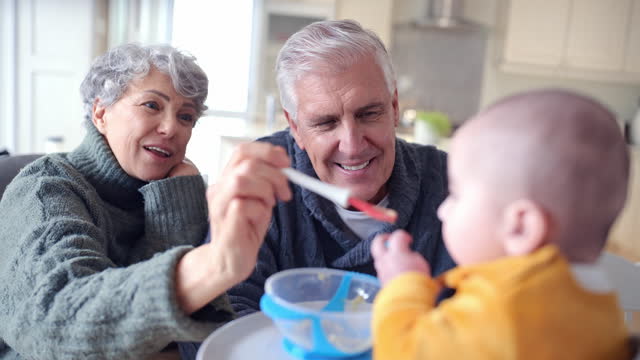 Baby, feeding food and breakfast with happy grandparents by table at home with porridge for nutrition and child growth. Family, kitchen and meal for wellness and hungry kid with a spoon in high chair