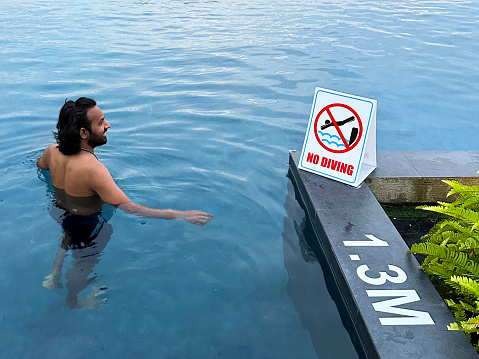 Stock photo showing close-up view of Indian man swimming, splashing and playing in infinity swimming pool with no diving warning sign for health and safety to prevent accidents and potential drowning hazard for swimmers.