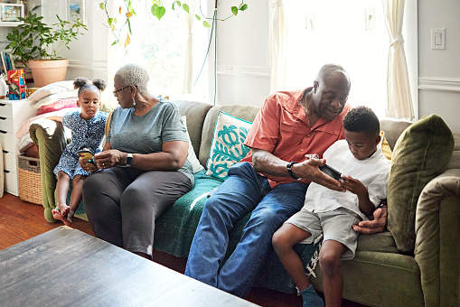 Senior Black couple sitting on sofa with grandchildren aged 4 and 7, using smart phone and playing video game in family home.