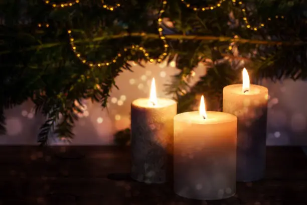 Three burning candles in front of dark fir branches with Christmas decoration and bokeh bubbles, seasonal holiday greeting card, copy space, selected focus, narrow depth of field